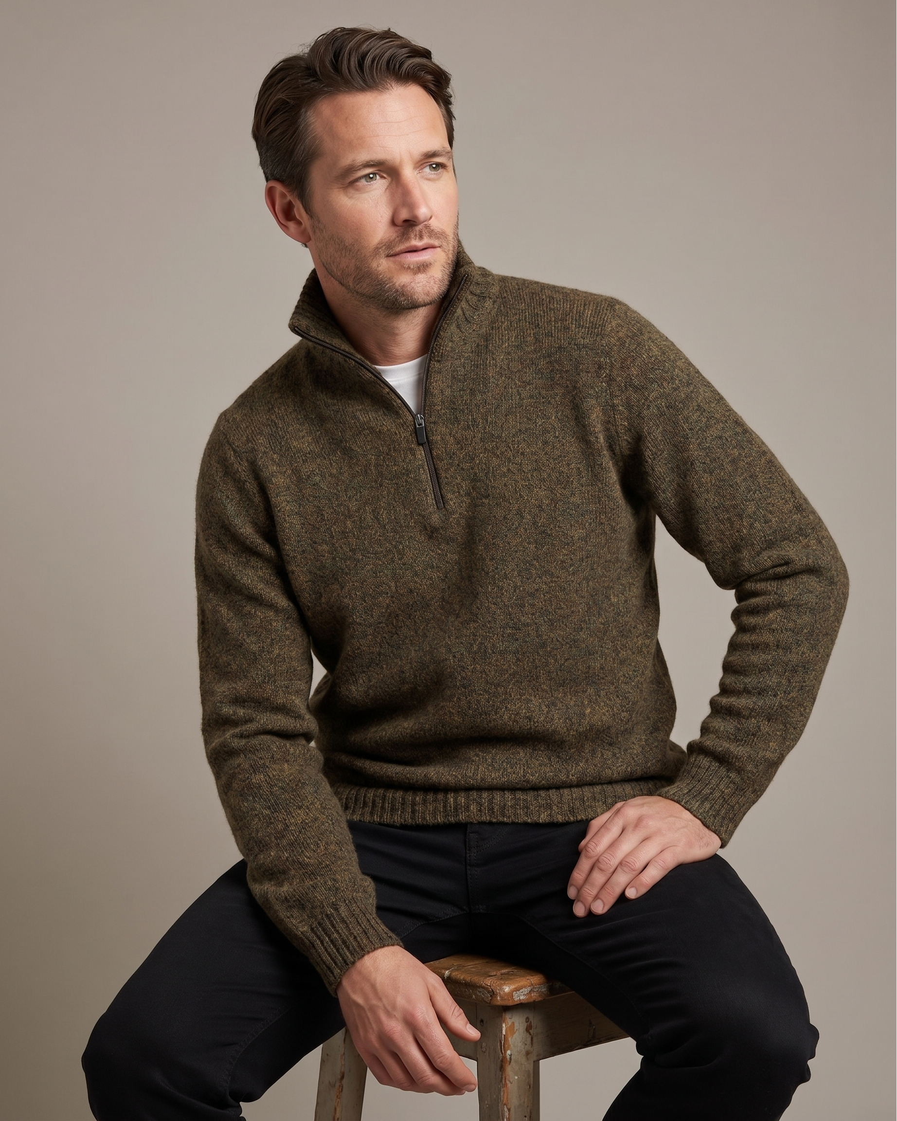 Man wearing a brown sweater sitting on a wooden stool against a beige background