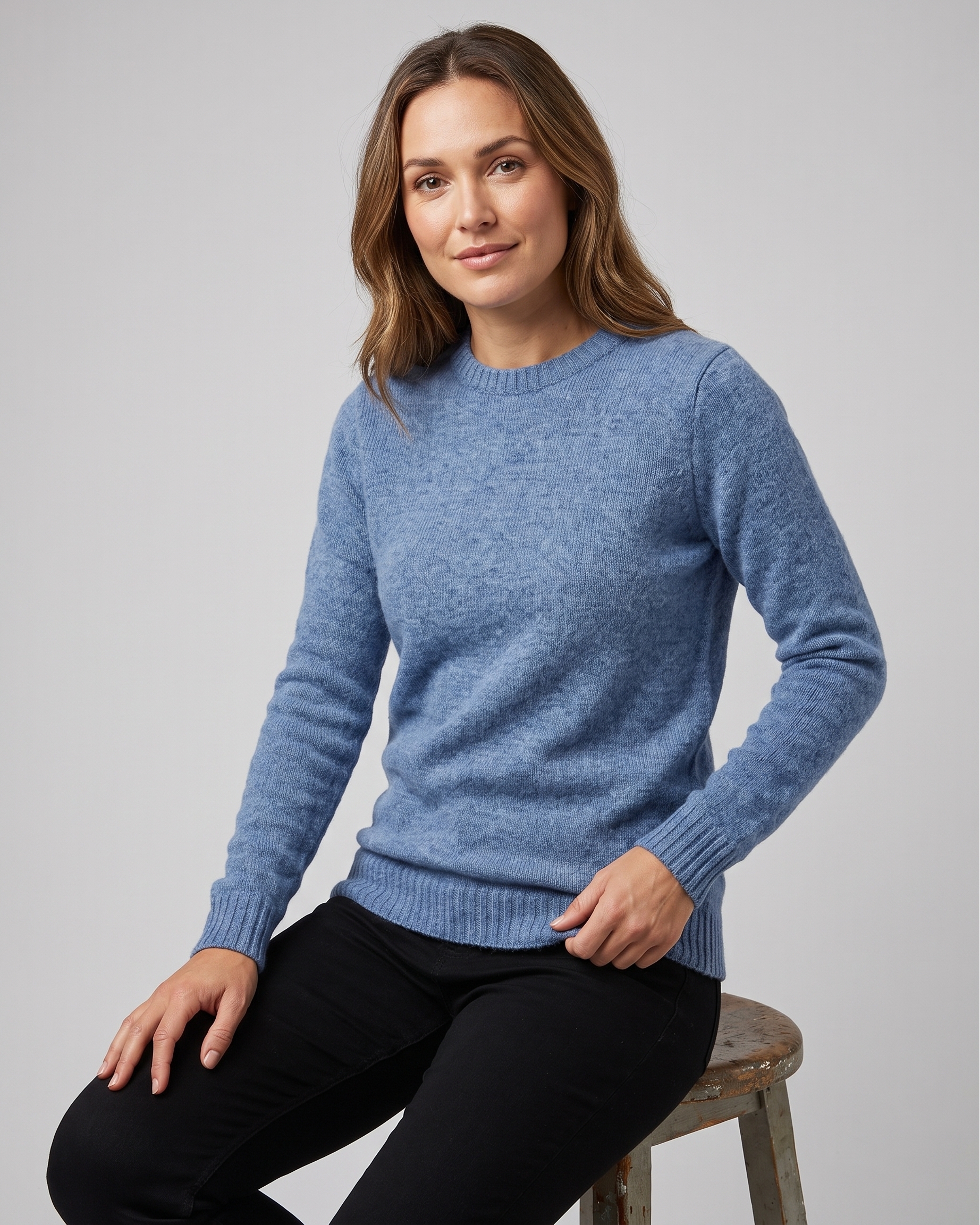 Woman wearing a blue sweater sitting on a stool against a gray background