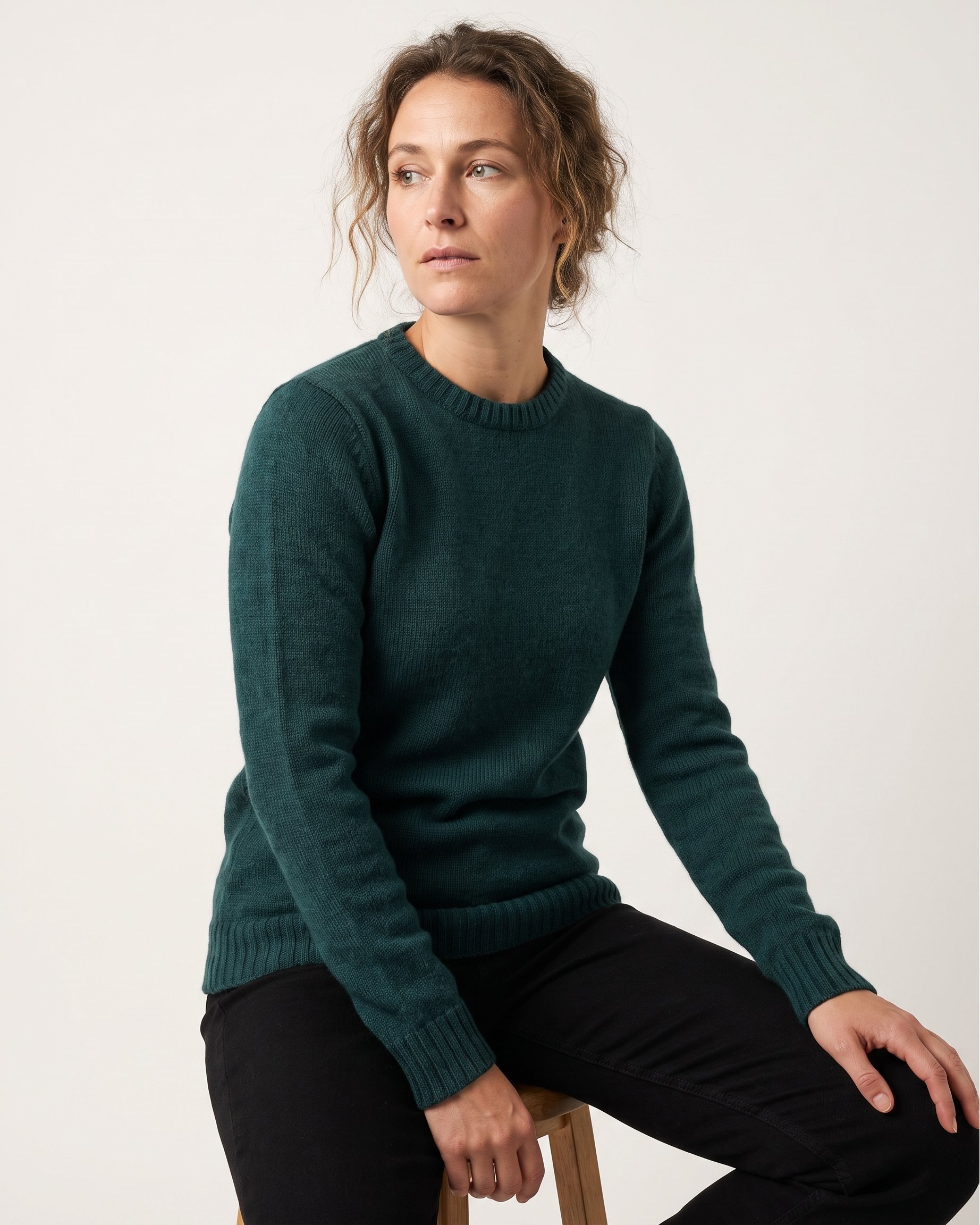 Woman wearing a teal sweater sitting on a stool against a white background
