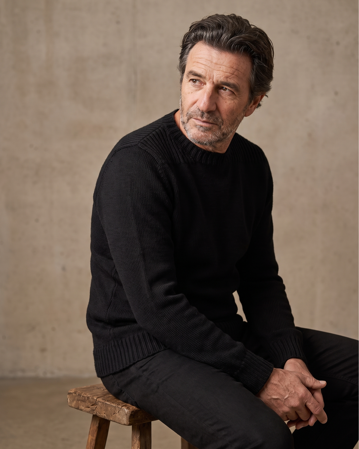 A man wearing a black ribbed knit sweater, seated on a rustic wooden stool against a textured beige concrete wall.