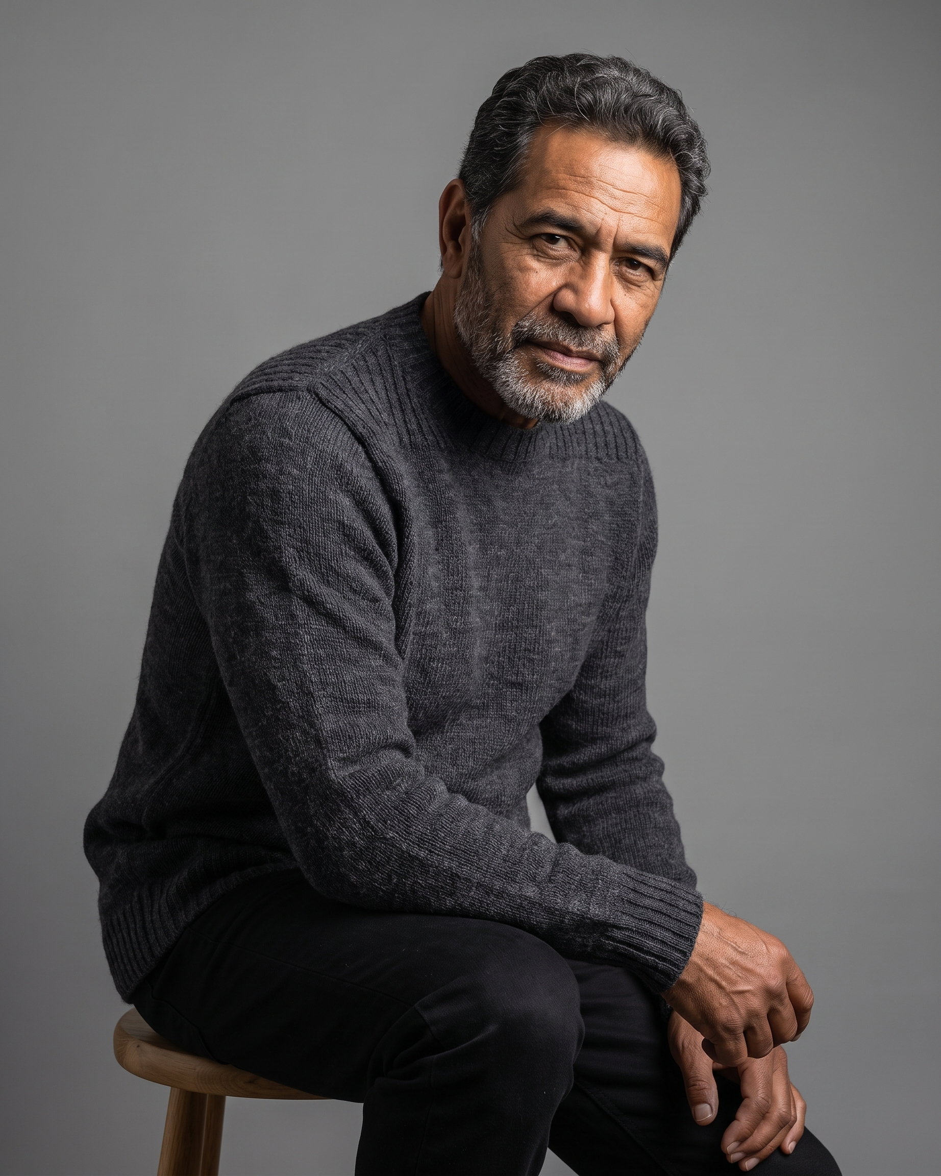 A man with graying hair and a beard sits on a wooden stool, wearing a dark gray knit sweater against a solid gray studio background.