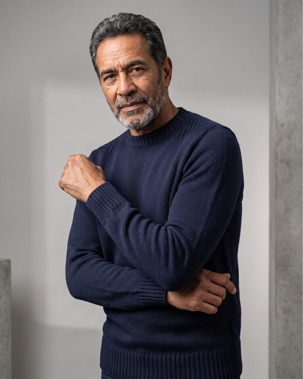 A man wearing a dark navy blue ribbed knit sweater, posing with his arms crossed against a neutral gray background.