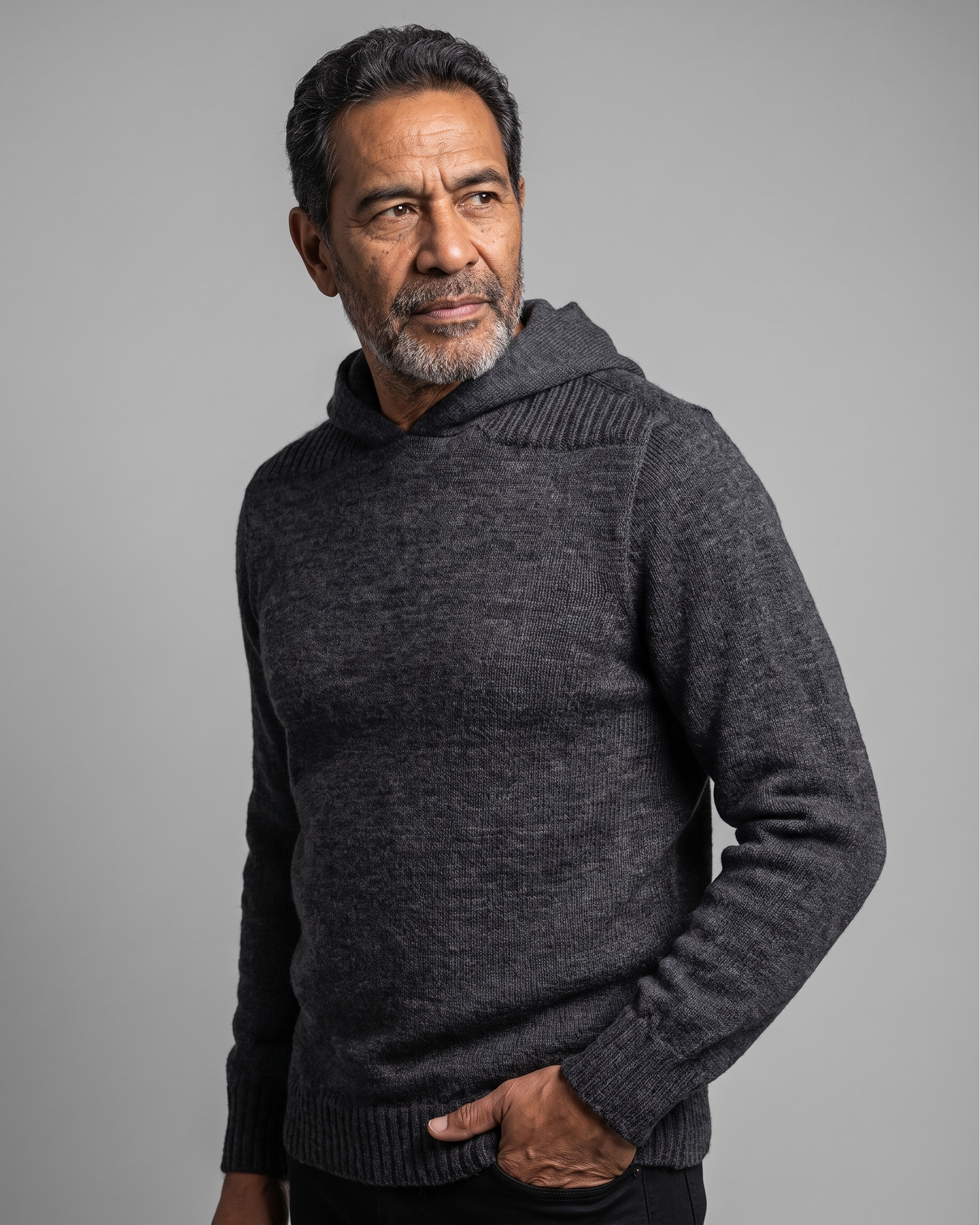 A man wearing a dark charcoal gray knit hooded sweater, standing against a solid gray studio background.