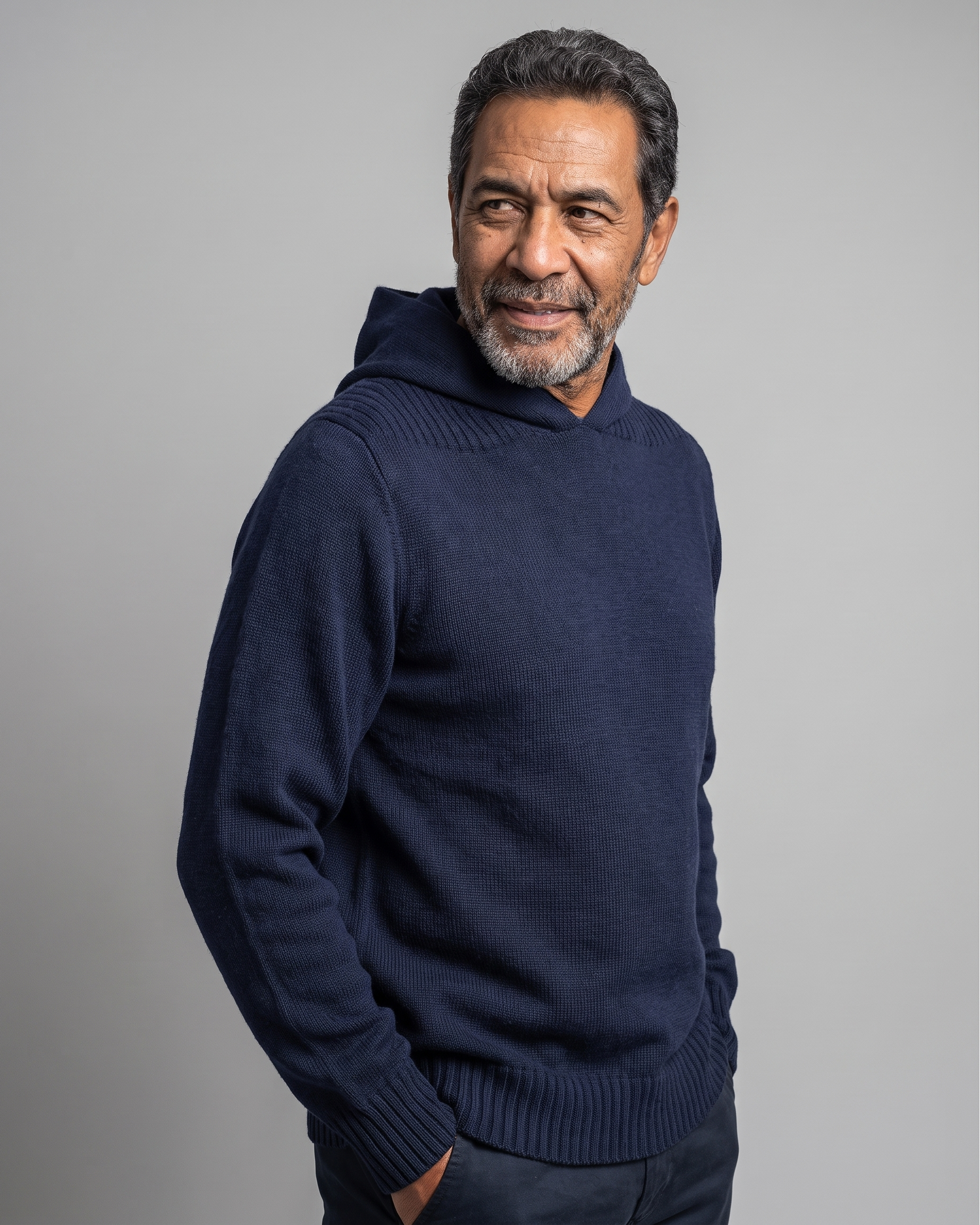 A man wearing a navy blue knit hooded sweater, standing against a solid gray studio background.