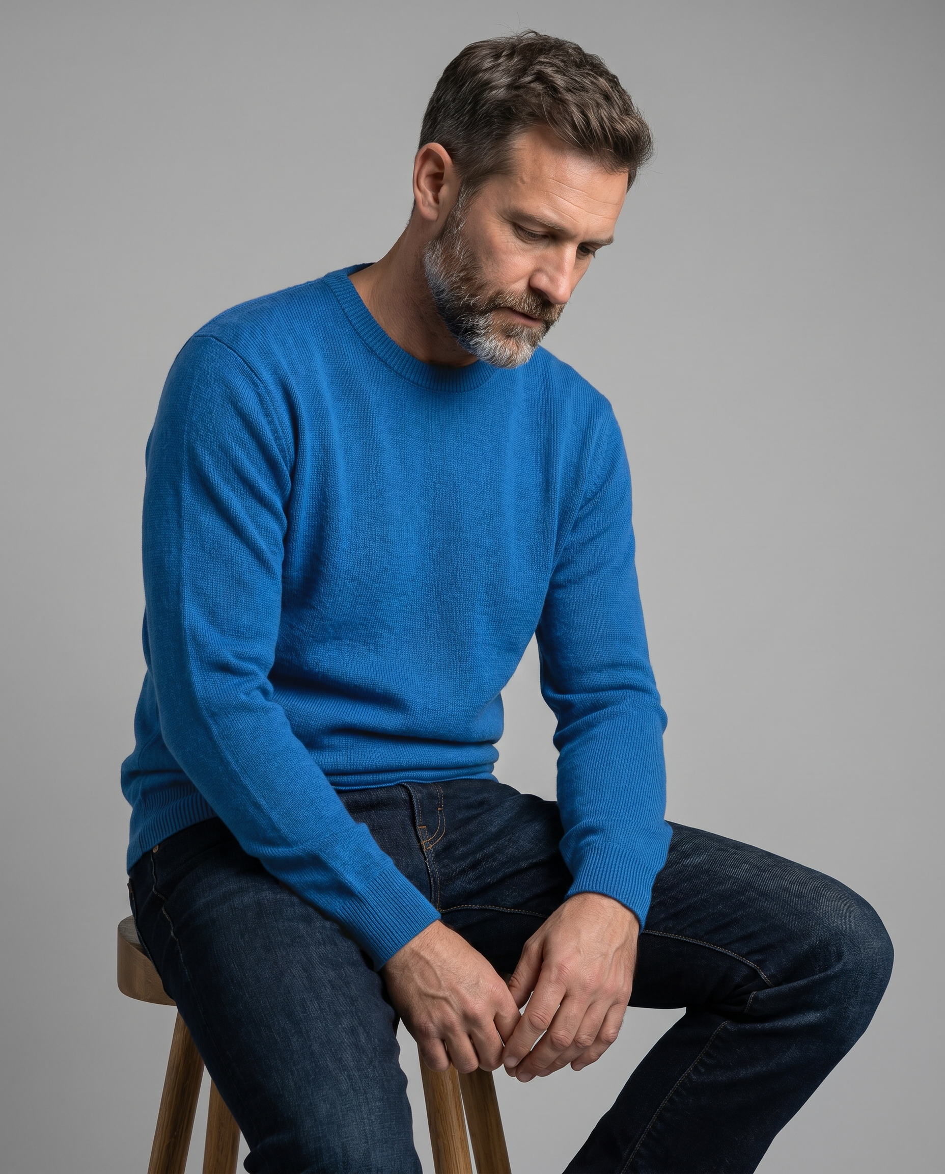 Man wearing a blue sweater and dark jeans sitting on a stool against a gray background