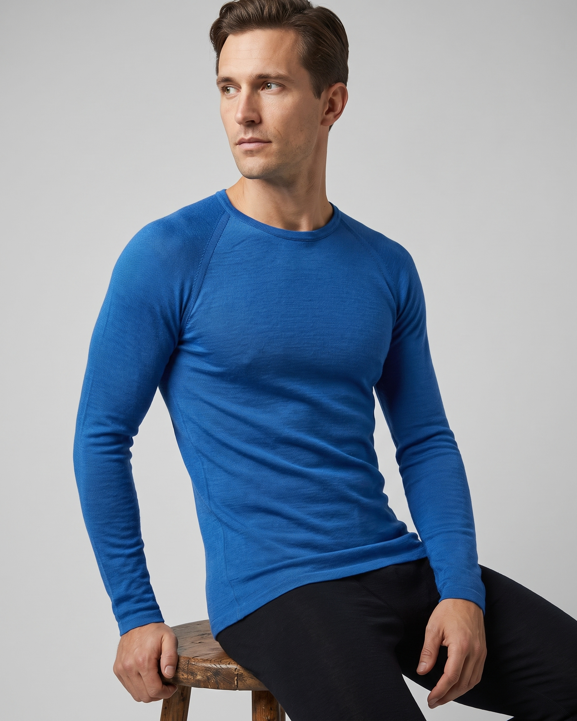 Man wearing a blue long-sleeve thermal sitting on a wooden stool against a gray background