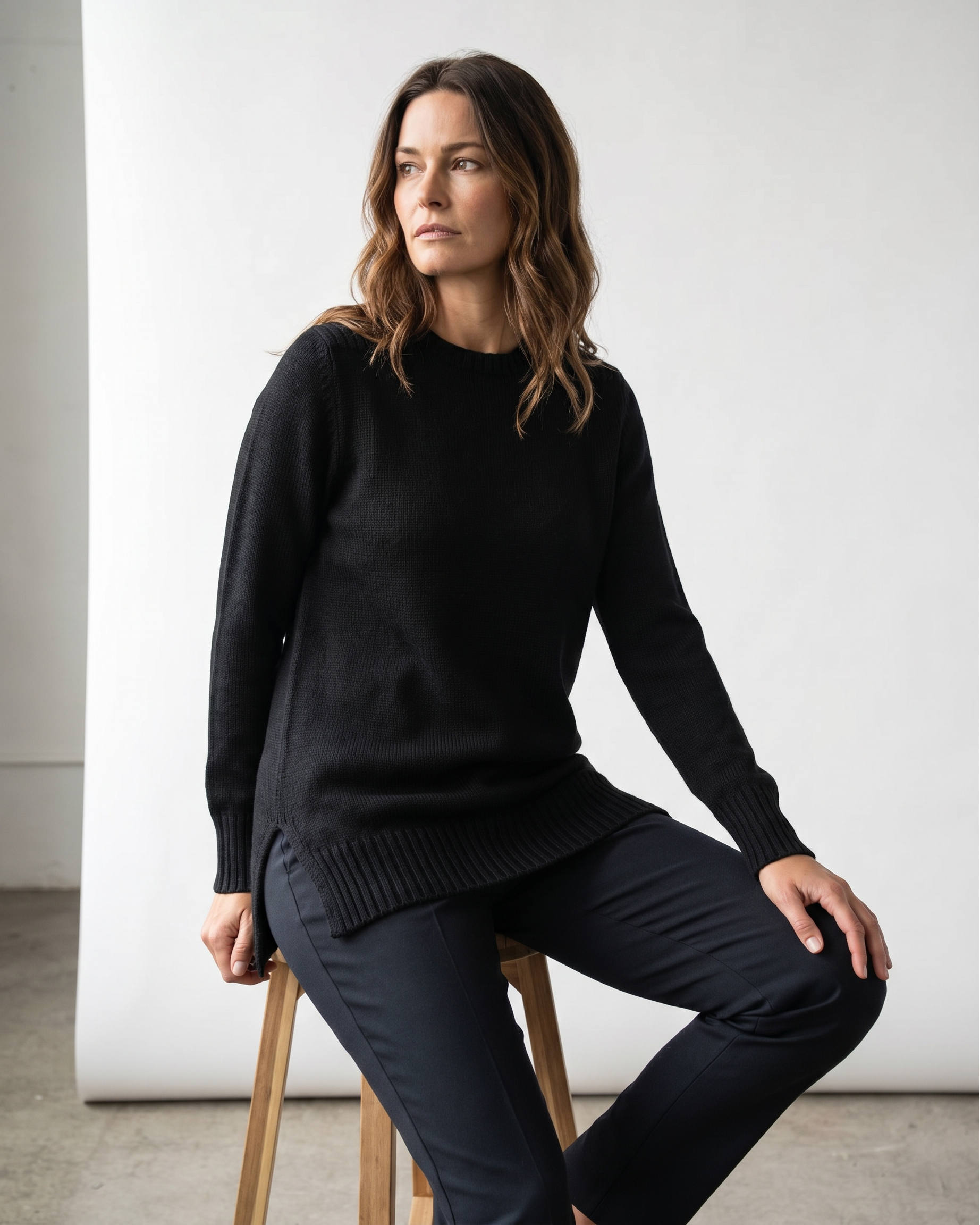 Woman wearing a black tunic sweater and dark pants sitting on a wooden stool against a white studio wall.