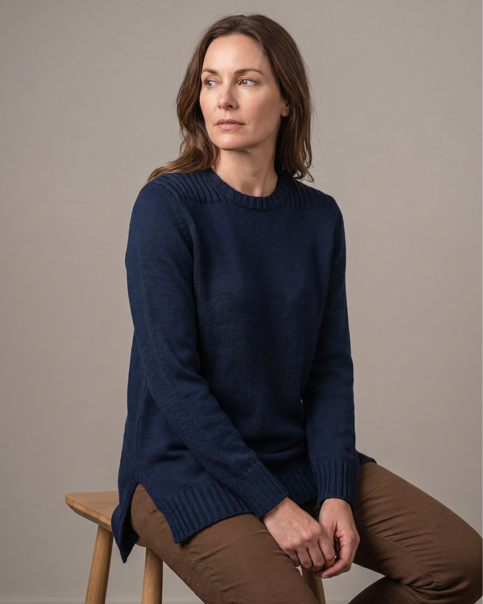 Woman wearing a navy tunic sweater sitting on a wooden stool against a plain background