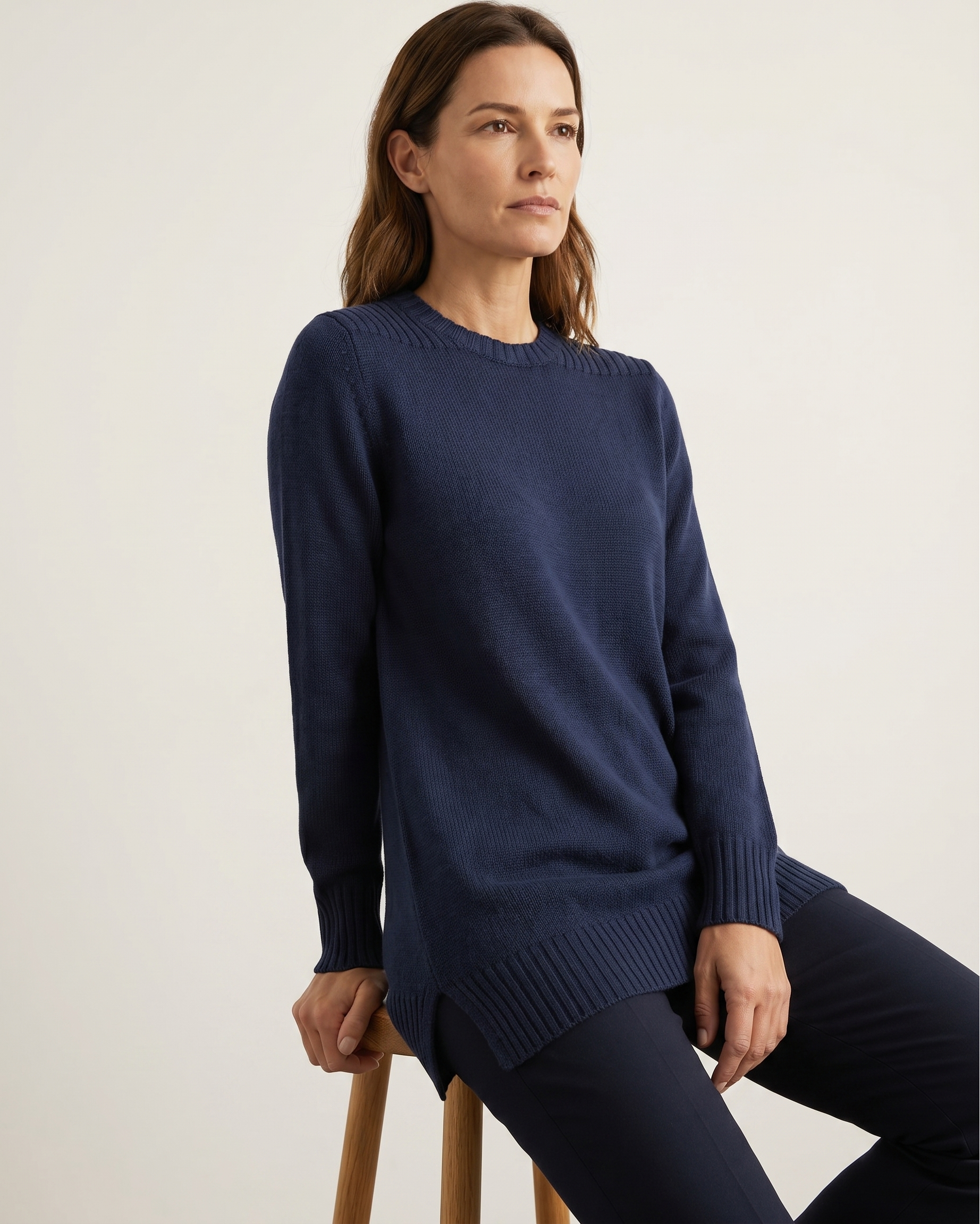 Woman wearing a navy blue tunic sweater sitting on a wooden stool against a plain background