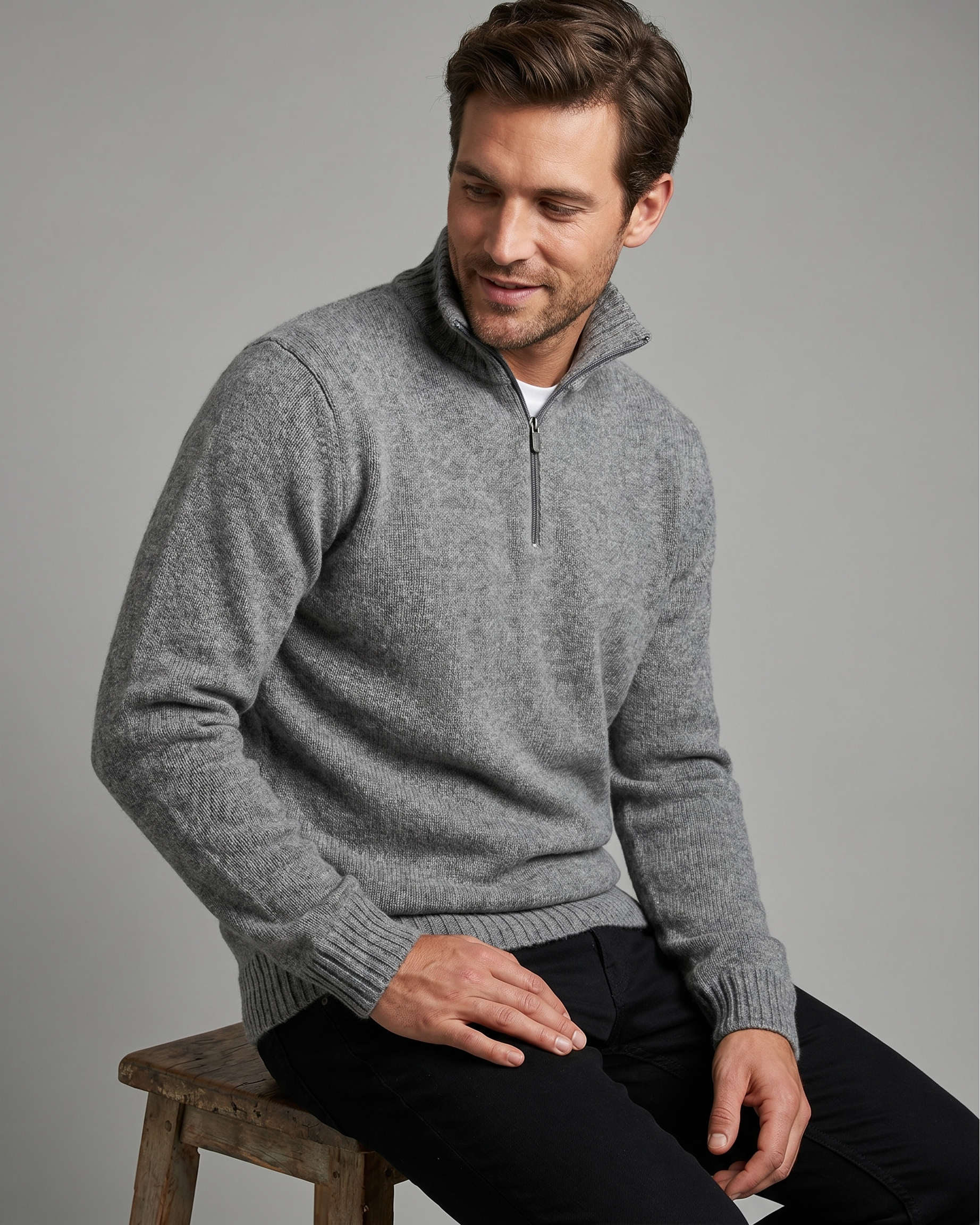 Man wearing a gray quarter-zip sweater sitting on a wooden stool against a gray background