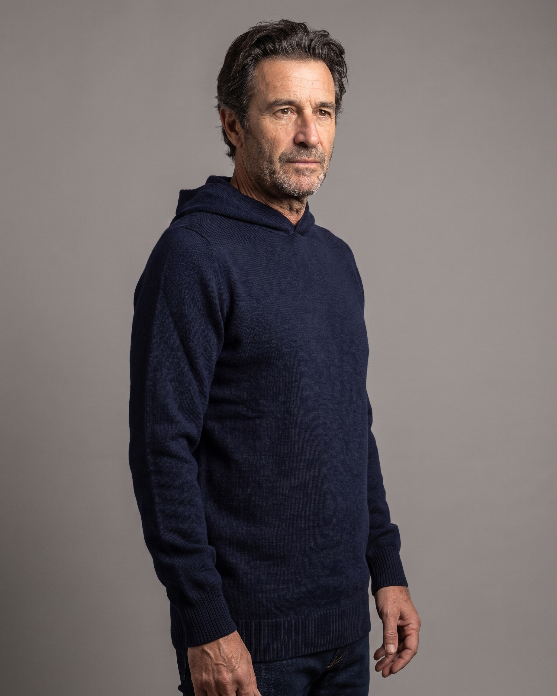 A man wearing a dark navy blue knit hoodie sweater, standing in profile against a solid gray studio background.