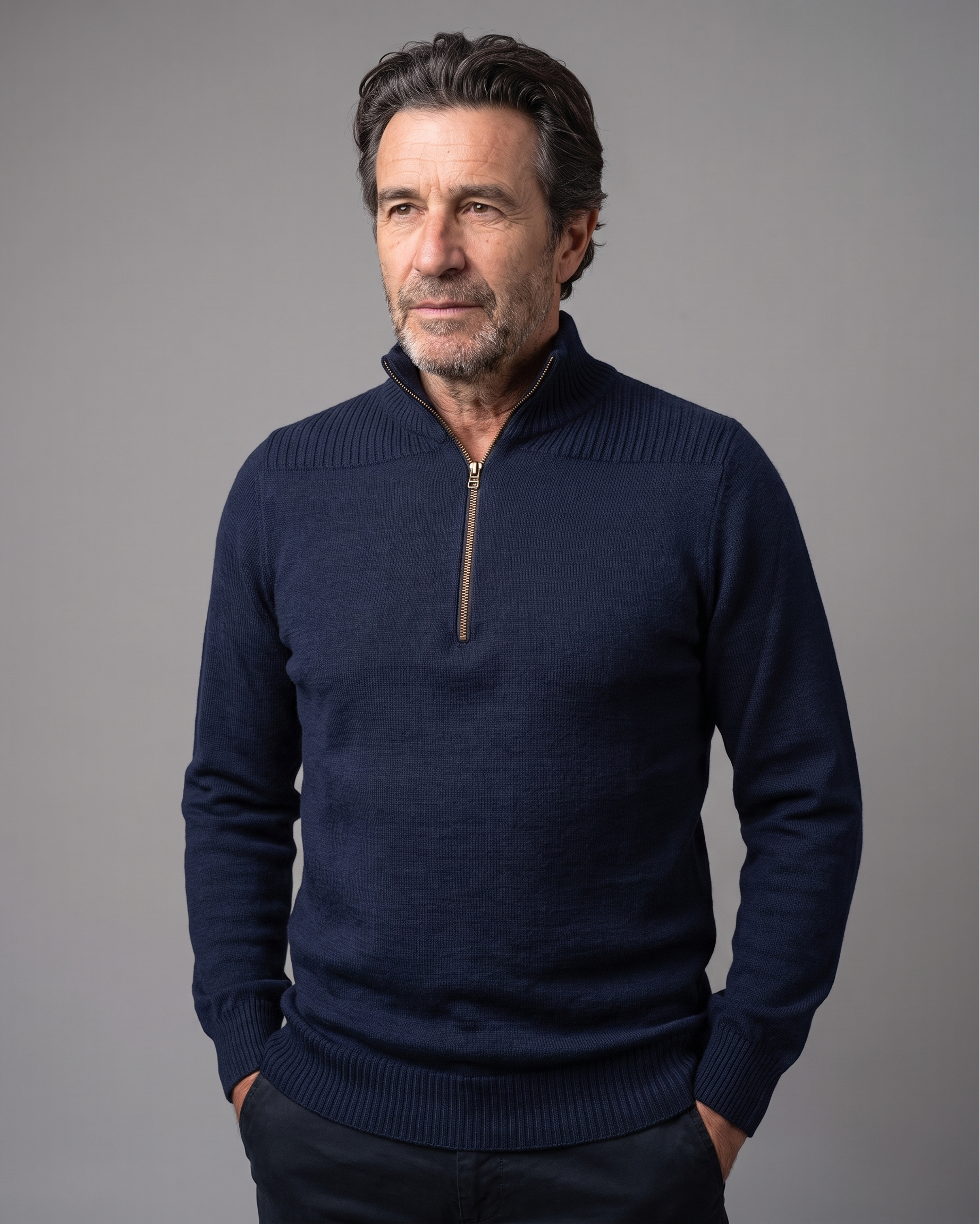 A man wearing a navy blue quarter-zip knit sweater with a ribbed mock neck, cuffs, and hem, standing against a solid gray studio background.
