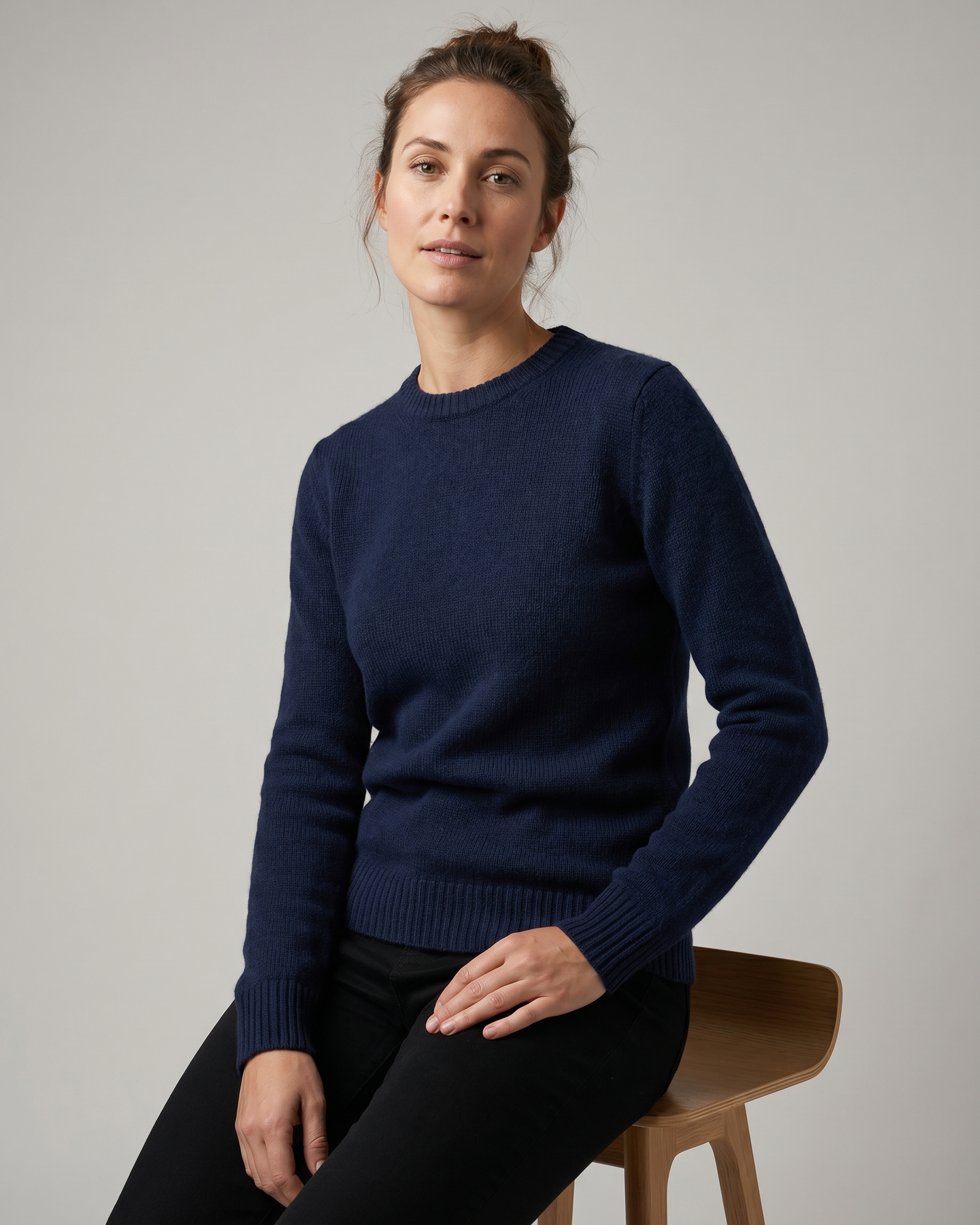Woman wearing a navy blue sweater sitting on a wooden stool against a plain background
