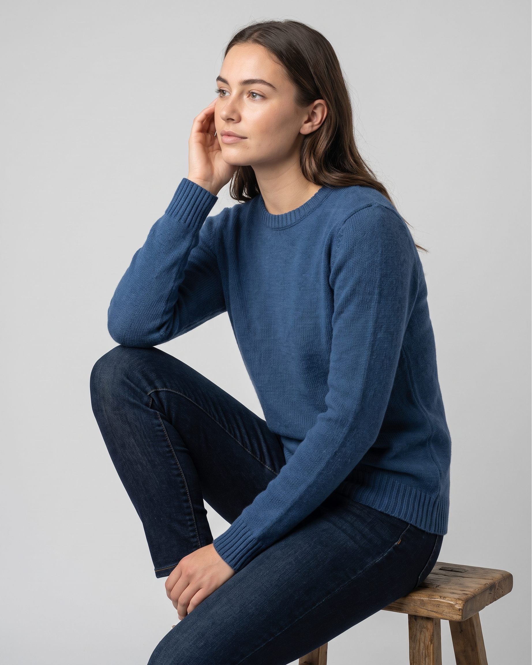 Woman wearing a pale blue sweater sitting on a wooden stool against a gray background