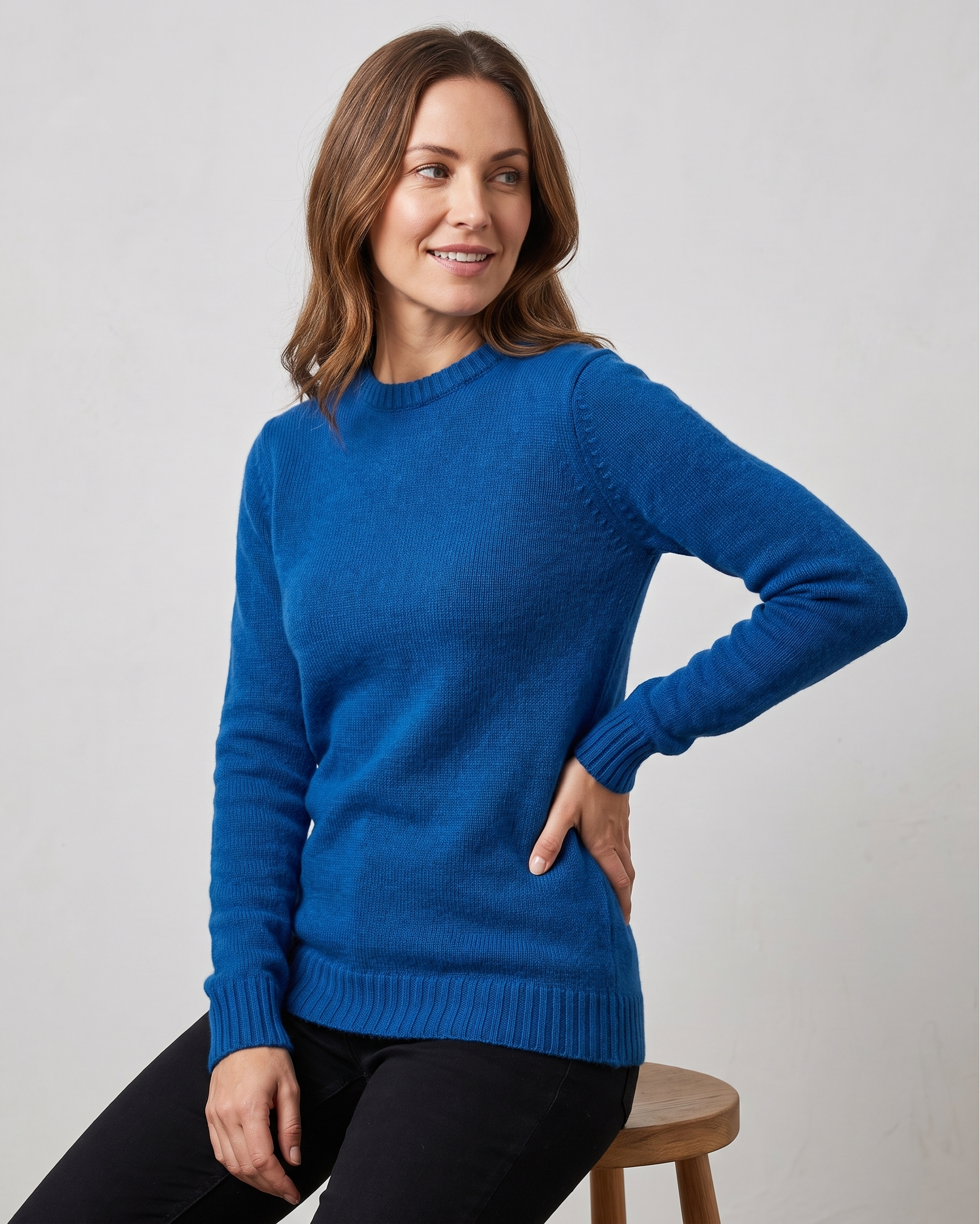 Woman wearing a blue sweater sitting on a wooden stool against a plain background