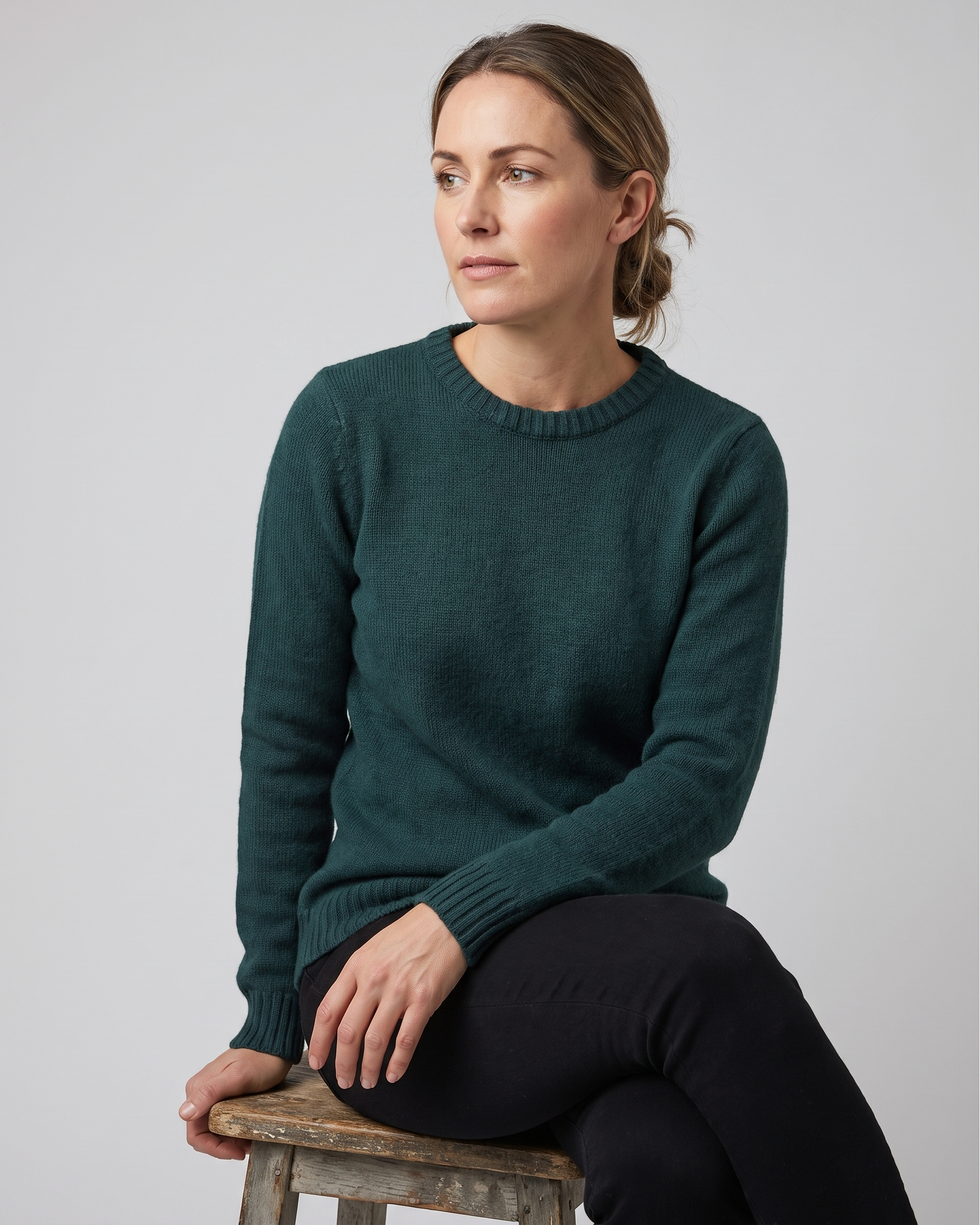 Woman wearing a teal sweater sitting on a wooden stool against a plain background