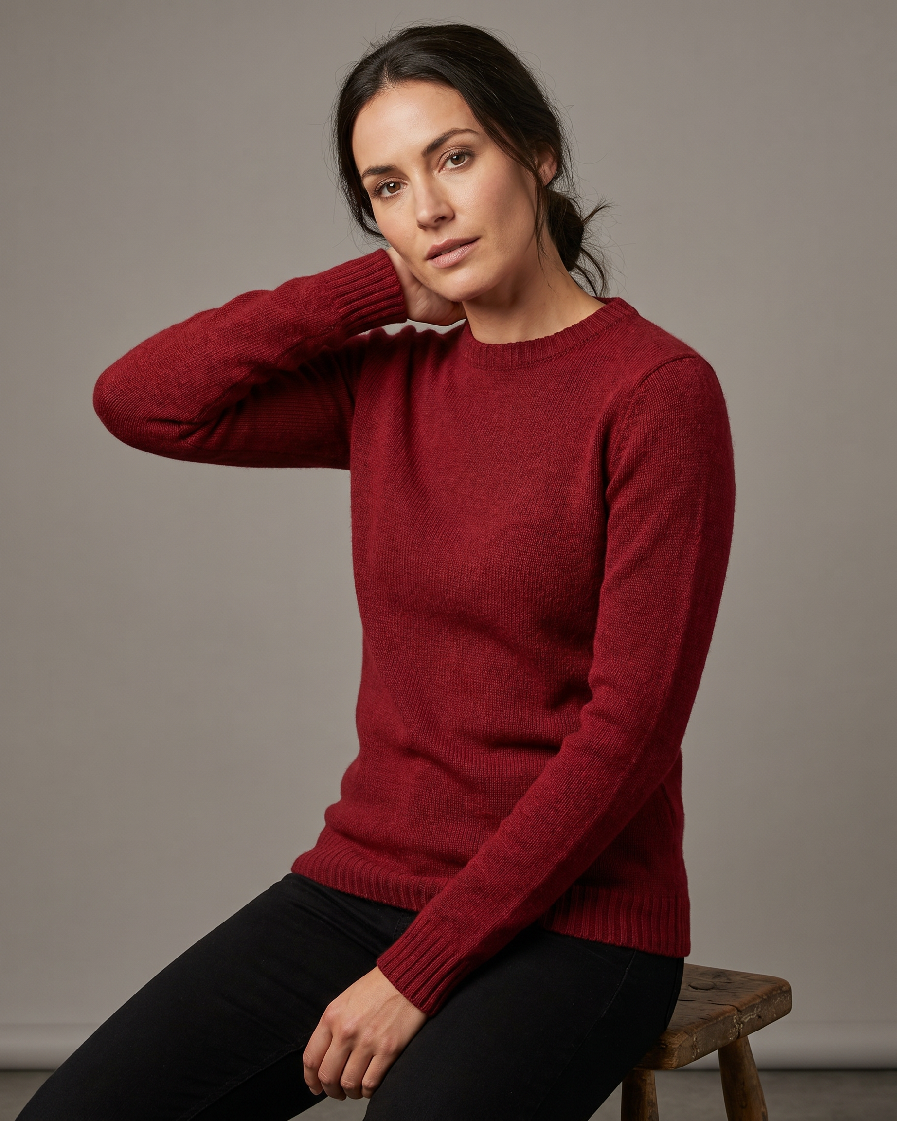 Woman wearing a red sweater sitting on a wooden stool against a plain background