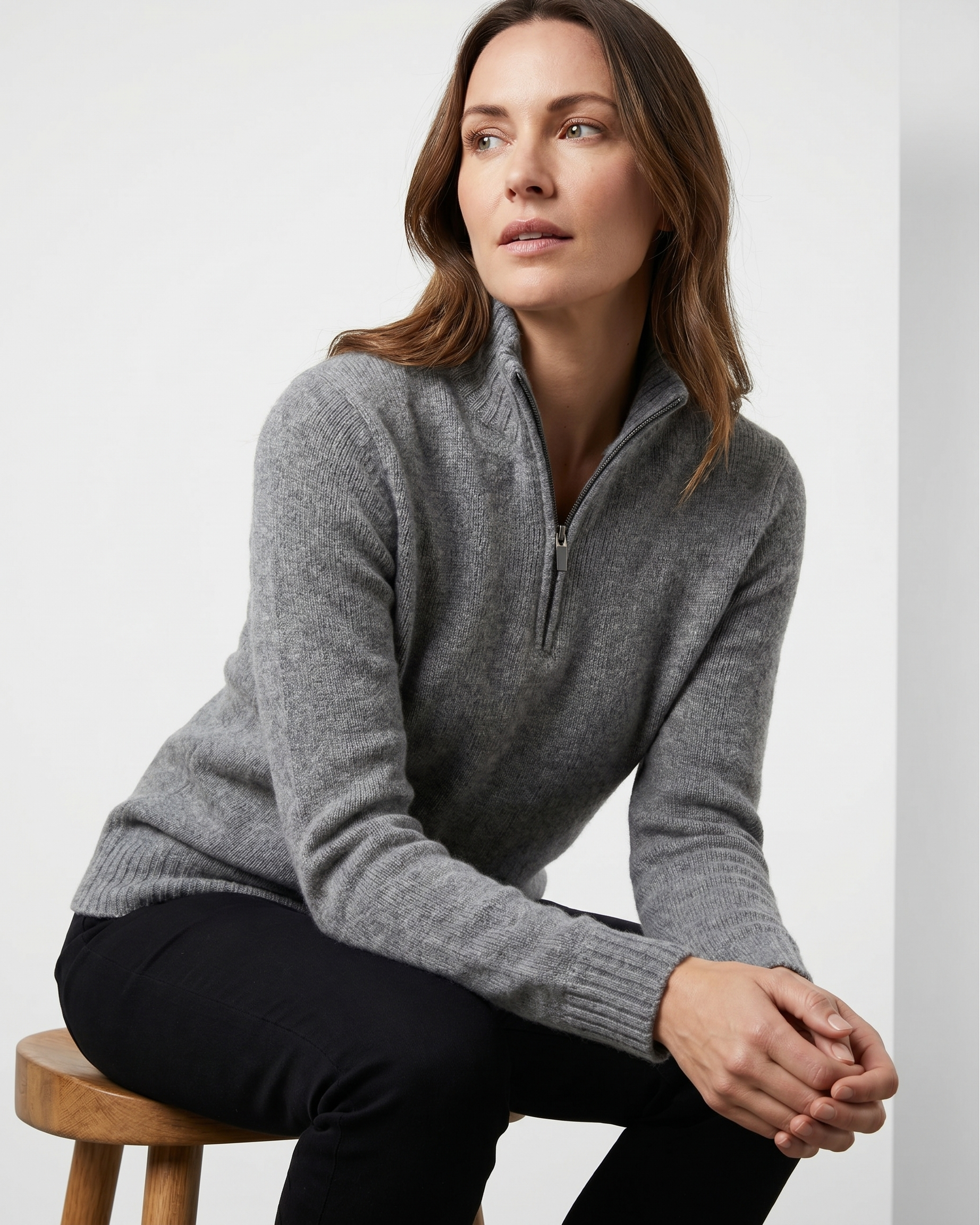 Woman wearing a gray sweater sitting on a wooden stool against a white background