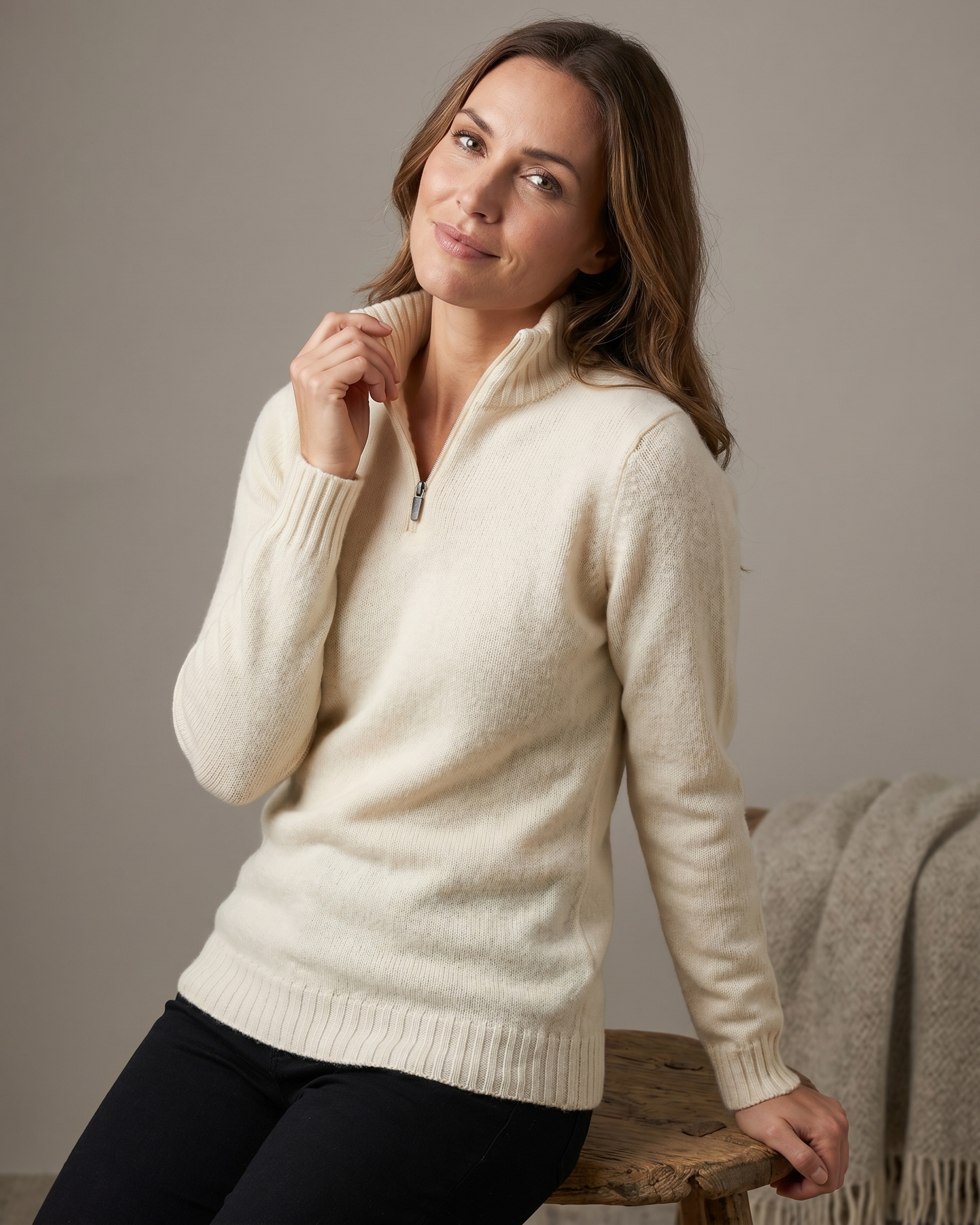 Woman wearing a cream sweater sitting on a wooden stool against a plain background