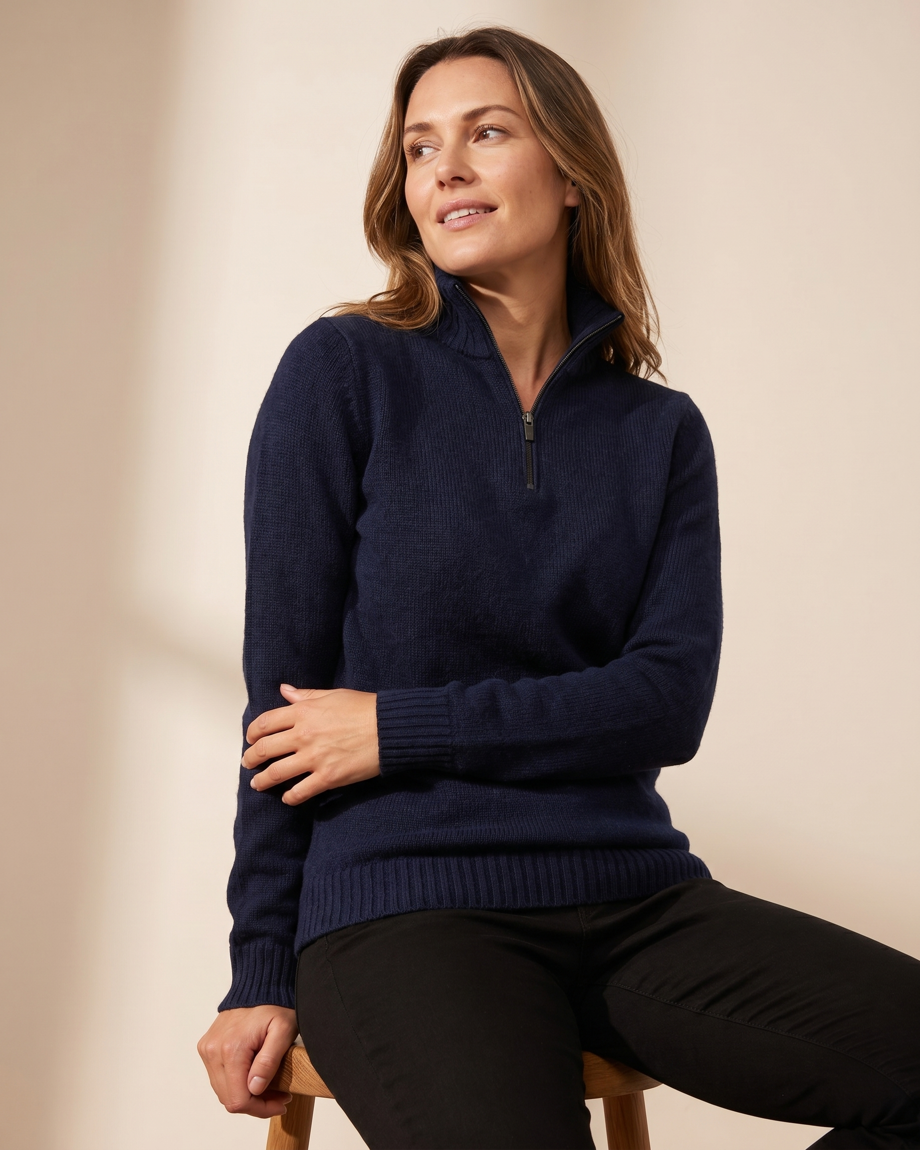 Woman wearing a navy blue sweater sitting on a wooden stool against a beige background