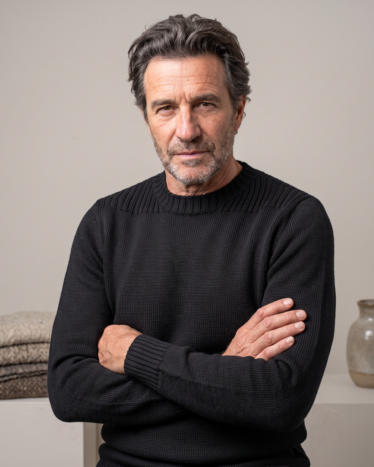 A man with gray hair and a beard wearing a black ribbed knit sweater, standing with arms crossed in front of a neutral beige background.
