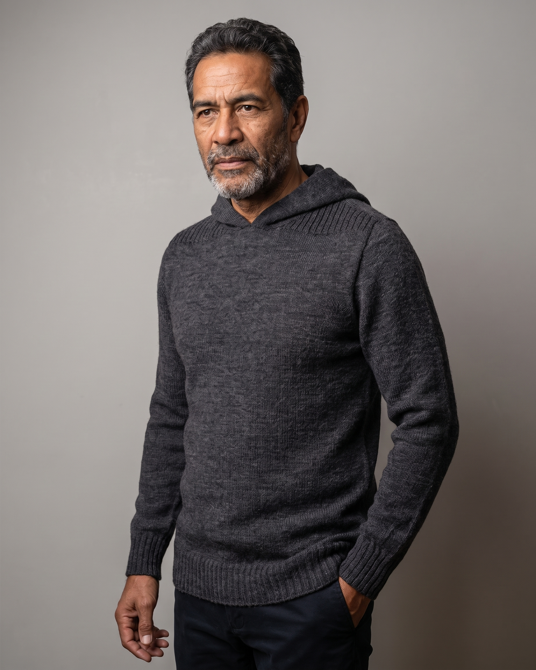 A man wearing a dark gray ribbed knit hooded sweater, standing against a neutral gray studio background.