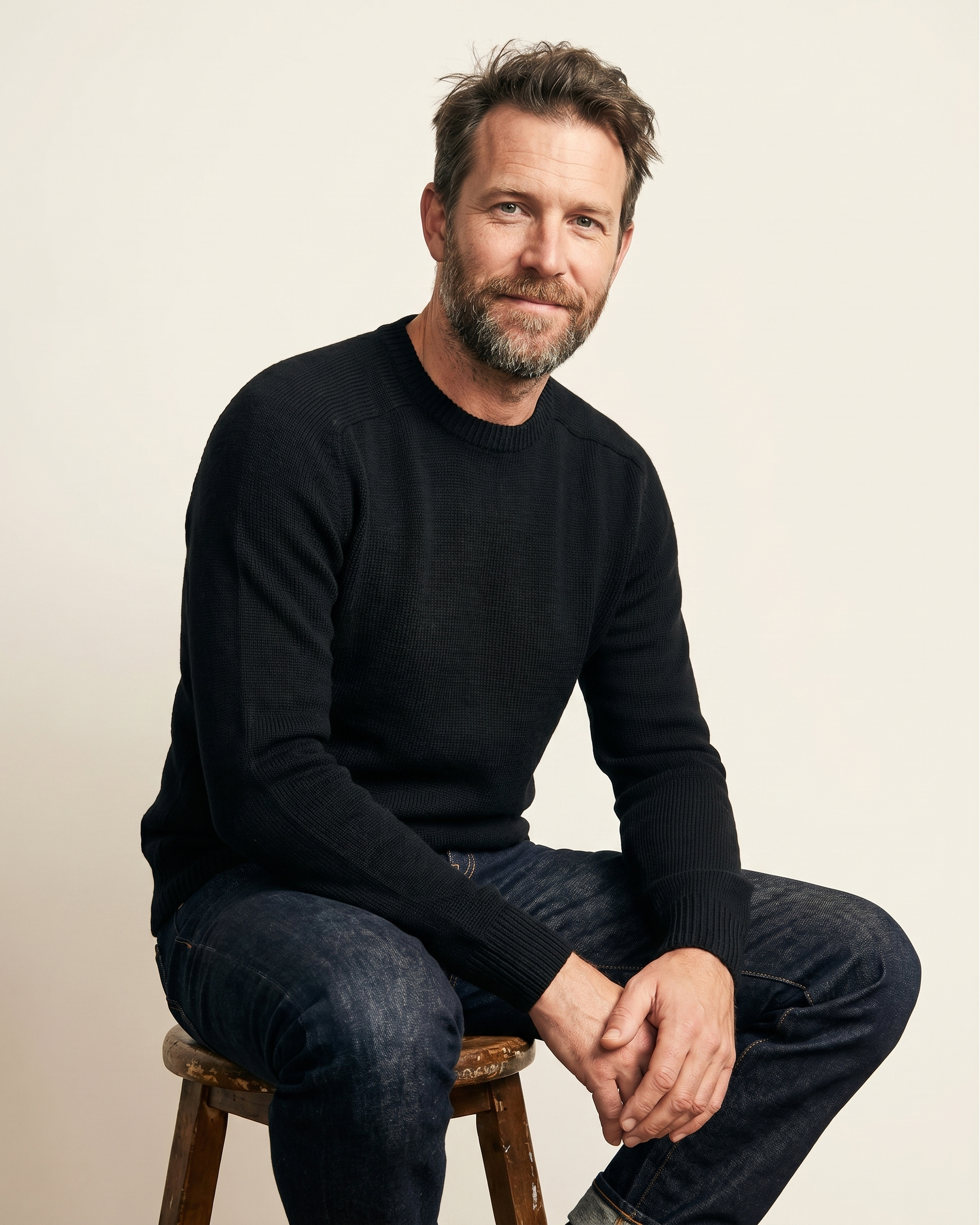 Man sitting on a stool wearing a black sweater and jeans against a plain background