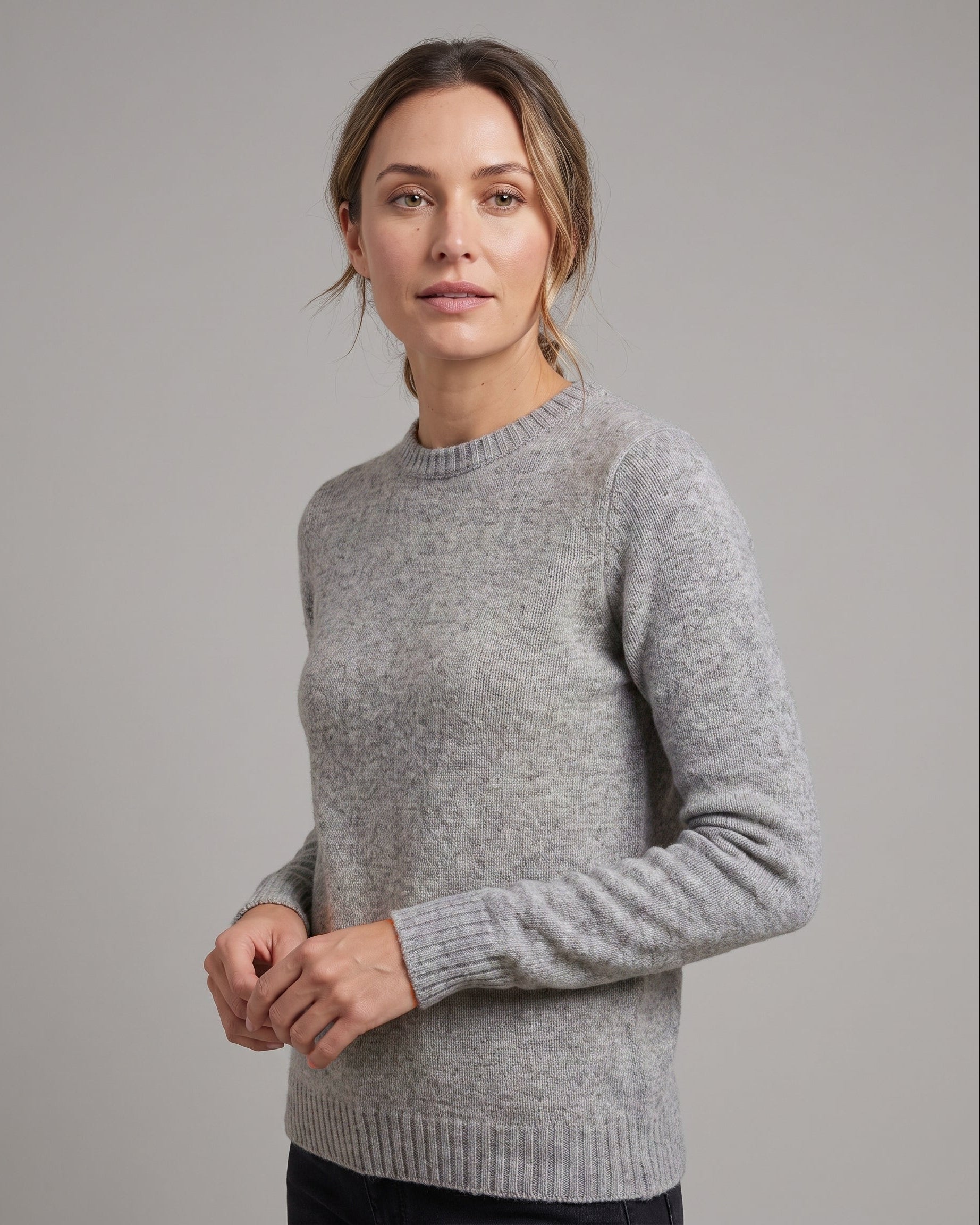 A woman wearing a light gray knit crewneck sweater, photographed against a solid gray studio background.